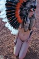 A naked woman wearing an indian headdress sitting on the ground.