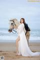 A woman in a white dress standing next to a white horse on the beach.